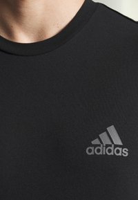 Mustat urheilupaidat, joka on valmistettu teksturoidusta kankaasta ja jossa on pyöreä kaula-aukko. Vasemmassa rintakappaleessa on harmaa Adidas-logo.