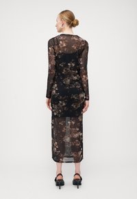 Robe noire transparente à manches longues, motif floral dans des tons bruns. Corsage ajusté menant à une jupe fluide et transparente.