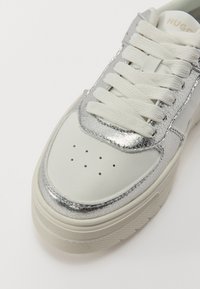 Baskets en cuir blanc avec des accents métalliques argentés, doublure texturée et lacets plats. Présente des perforations sur le bout pour une bonne respirabilité.
