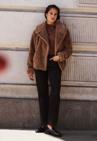 Bruine nepbontjas met brede revers, gedragen over een bruin luipaardprinttop, gecombineerd met zwarte rechttoe-rechtaan broek en glanzende zwarte loafers.