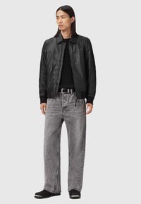 Man med långt hår, klädd i en svart läderjacka, svart tröja, grå vida jeans, svart bälte och svarta skor, stående mot en grå bakgrund.