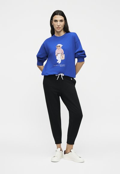 Γυναίκα που φοράει μπλε φούτερ με γραφικό αρκούδας, μαύρα αθλητικά παντελόνια και λευκά αθλητικά παπούτσια, στέκεται με τα χέρια πίσω από την πλάτη σε λευκό φόντο.