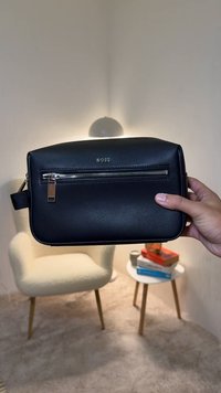 Μαύρη δερμάτινη τσάντα με ορθογώνιο σχήμα, που διαθέτει μπροστινή τσέπη με φερμουάρ, λεία υφή και χρυσές λεπτομέρειες. Εμφανίζεται το λογότυπο της μάρκας.