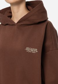 Felpa con cappuccio marrone realizzata in tessuto morbido, caratterizzata da un piccolo logo ricamato in beige chiaro nella zona sinistra del petto.