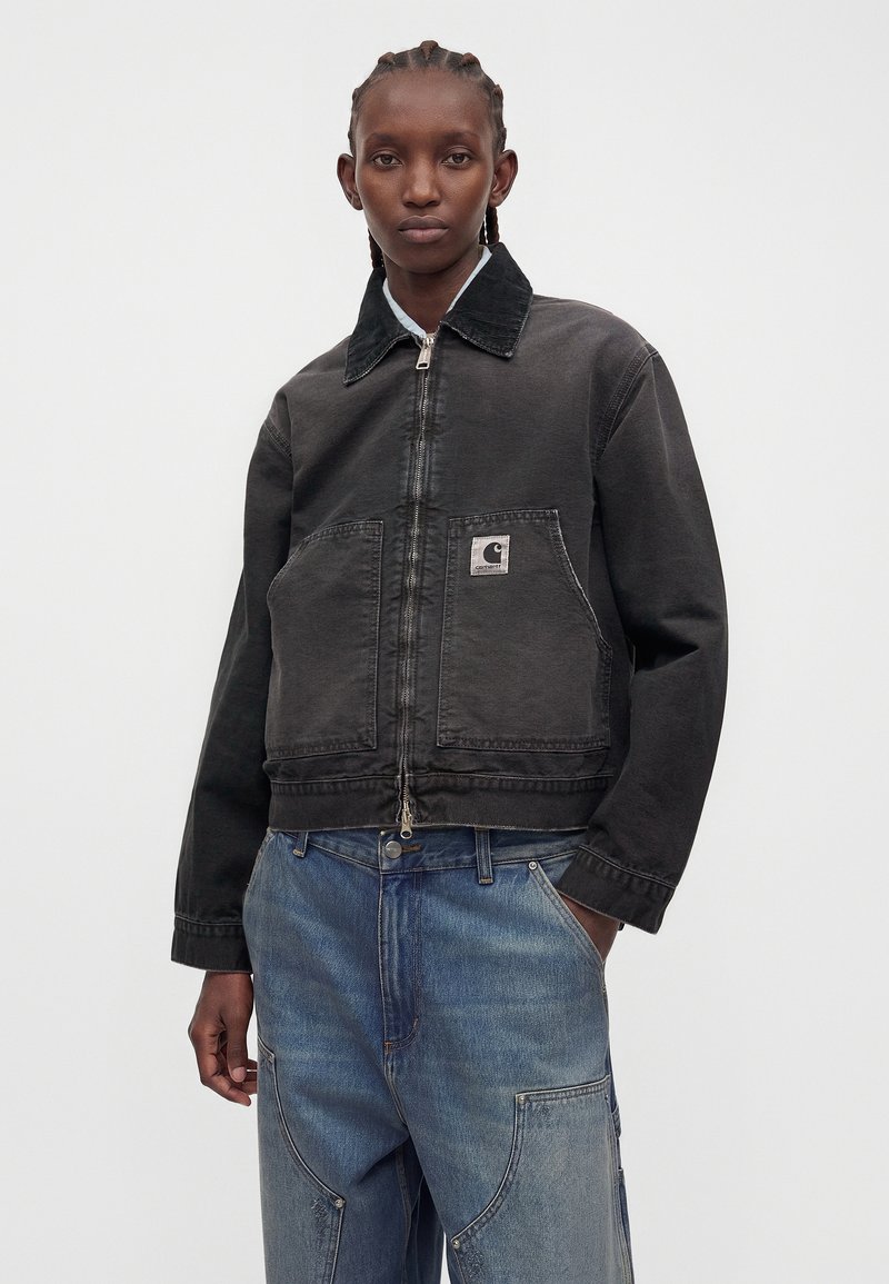 Personne portant une veste zippée noire avec le logo Carhartt et un jean bleu ample, debout devant un fond blanc uni.
