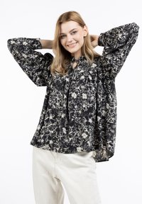 Black floral patterned blouse with billowy sleeves, a tie neck, and flowing design. Paired with light beige pants for contrast.