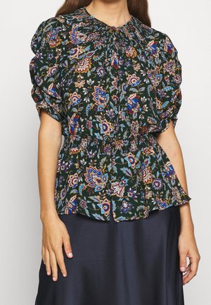 Vrouw draagt een donkergroene bloemenblouse met pofmouwen, geplooide halslijn en gerimpelde taille, gecombineerd met een donkere rok.
