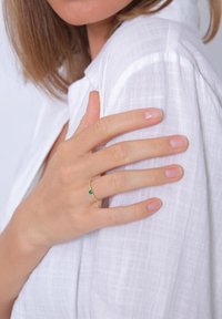 Goldring mit einem kleinen grünen Edelstein, getragen an einer Hand, die auf einem weißen strukturierten Stoff ruht. Sauberer Nagellack auf den Fingern.