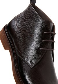 Botte en cuir marron au fini texturé, avec un bout arrondi, des coutures discrètes et trois œillets pour les lacets. Semelle en caoutchouc marron.