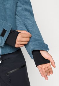 Modrá kordurová bunda s texturovaným povrchem. Obsahuje černý vnitřní rukáv se otvorem na palec. Rukáv lze nastavit ručně pomocí suchého zipu.