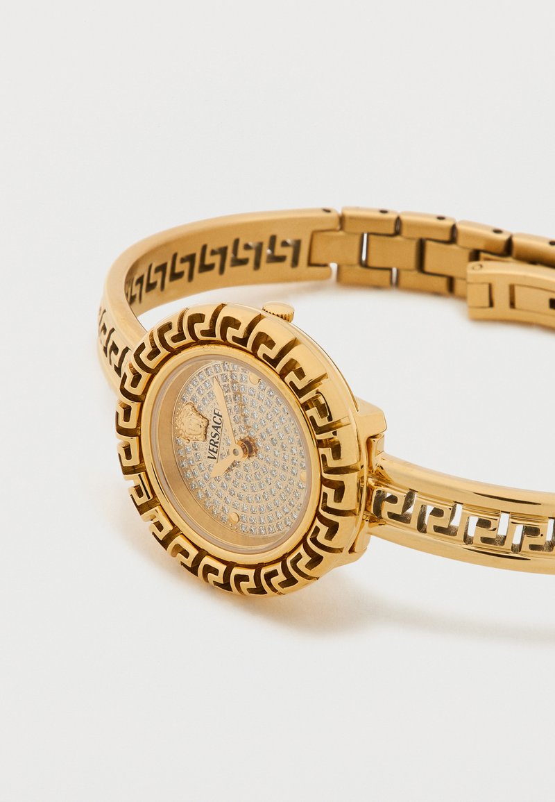 Goudkleurig horloge met een ronde wijzerplaat, bezet met kristallen, voorzien van een patroonband en het Versace-logo. Het patroon bevat details van een Griekse sleutel.
