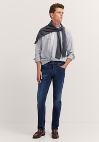 Hellblau gestreiftes Hemd, dunkelgrauer Pullover über den Schultern, blaue Jeans und braune Lederschuhe. Einfaches und klassisches Design.