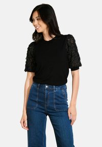 Zwarte top met gestructureerde kantmouwen, gecombineerd met hoog-taille blauwe denim jeans. De mouwen hebben een bloemenpatroon en een geplooid ontwerp.