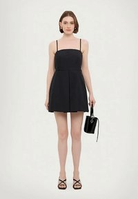 Sieviete, tērpta melnā bez piedurknēm mini kleitā un melnās sandales, tur mazu melnu rokassomu pret vienkāršu baltu fona.