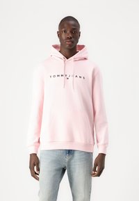 Hanorac roz deschis cu șnururi, buzunar mare în față și logo-ul negru "TOMMY JEANS". Asortat cu blugi din denim decolorat.