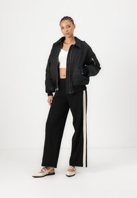 Veste bomber noire avec fermeture éclair et poignets côtelés, portée sur un haut court blanc et un pantalon noir avec des bandes blanches sur les côtés.