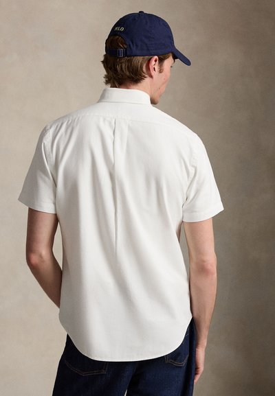 Férfi fehér rövid ujjú, gombos ingben és sötétkék Polo baseballsapkában, háttal egy semleges háttér előtt.