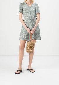 Kobieta w miętowej sukience z krótkim rękawem w białe kropki, trzymająca plecioną torbę-bucket, w czarnych płaskich sandałach na białym tle.