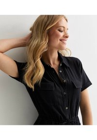 Vestido corto de manga corta negro con botones frontales, bolsillos en el pecho y cintura ajustable. La tela texturada y el corte relajado mejoran el diseño general.