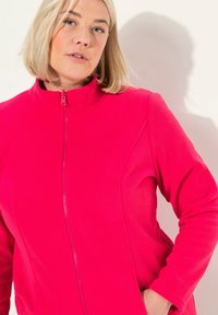 Fleece bunda v živé růžové barvě s vysokým límcem a plným zipem. Disponuje hladkou texturou a bočními kapsami pro praktičnost.