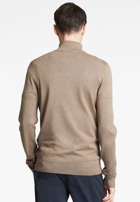 Beige strikkegenser med høy krage, lange ermer og ribbede mansjetter. Glatt tekstur, tettsittende design og sømløs konstruksjon er synlig.
