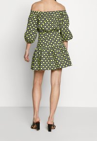 Schulterfreies Kleid mit puffigen Ärmeln, schwarz-grünes geometrisches Muster, taillierter Schnitt und Rüschensaum. Kombiniert mit schwarzen, hohen Sandalen.