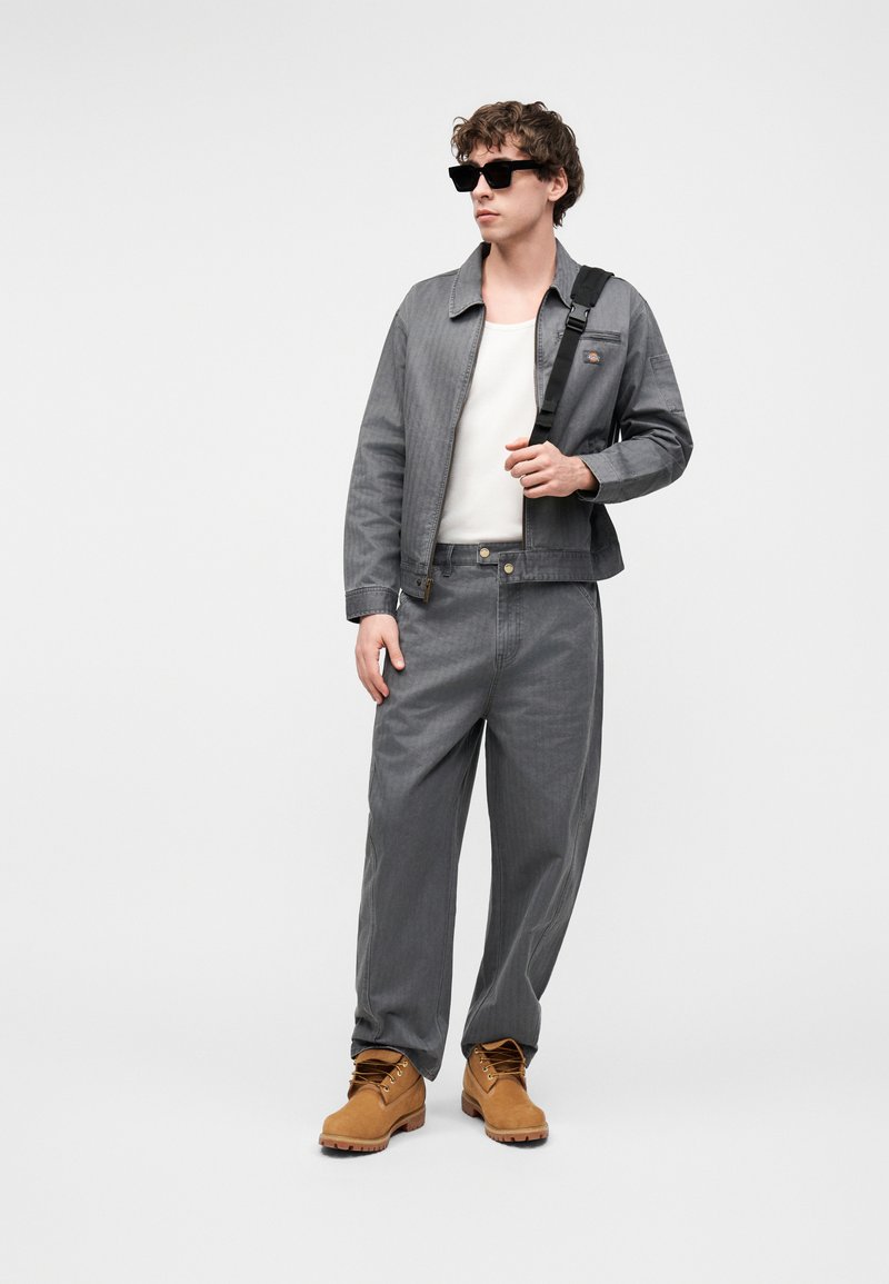 Jonge man die een grijs spijkerjack en broek draagt, wit overhemd, beige laarzen, zwarte zonnebril en een zwarte schoudertas.