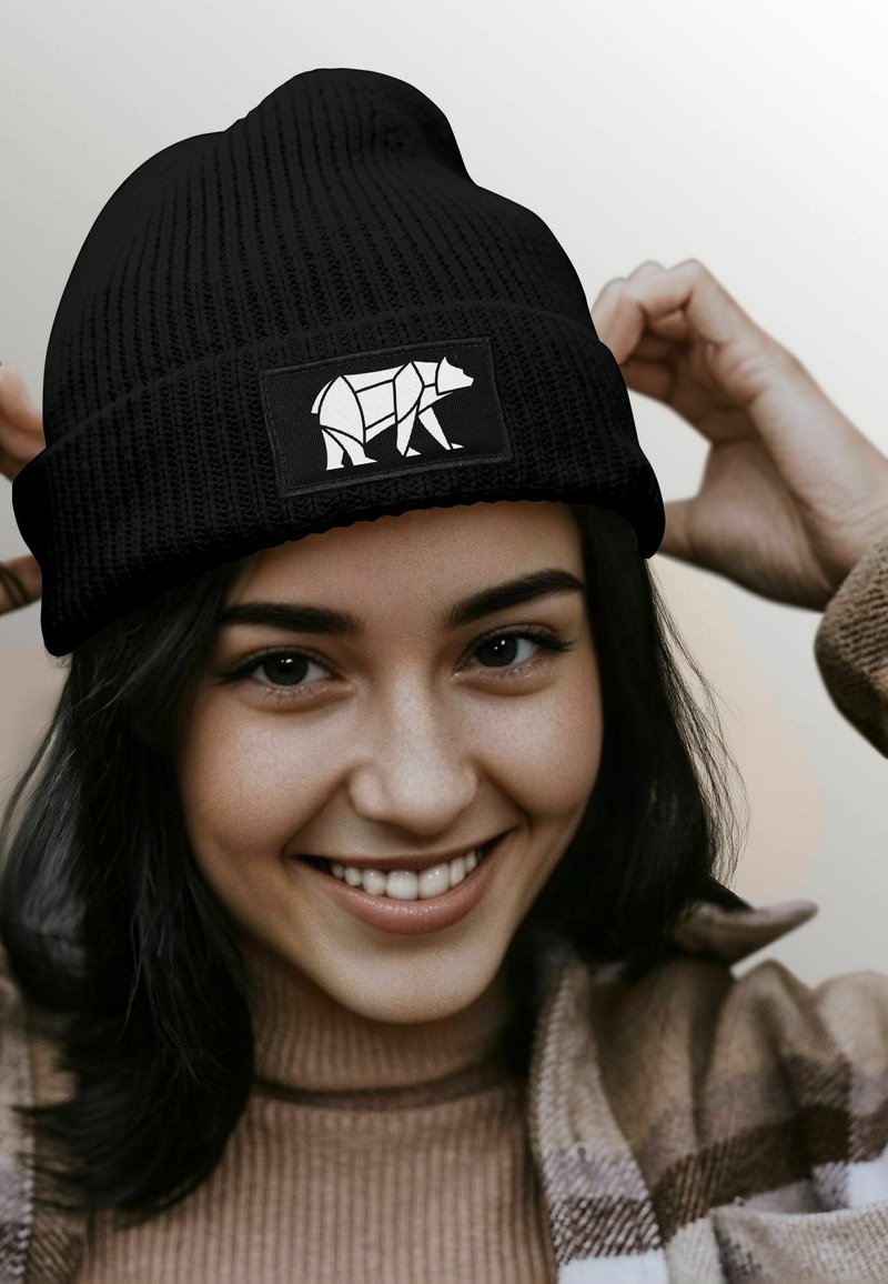 Black knit beanie with a ribbed texture and a white bear patch on the front. Model adjusts the beanie, smiling, wearing a beige sweater.