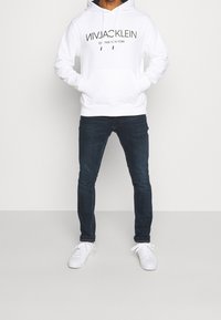 Valkoinen huppari mustalla tekstillä, kengurupocketilla ja nyöreillä. Yhdistetty tummansinisiin farkkuihin ja valkoisiin lenkkareihin. Minimalistinen muotoilu.