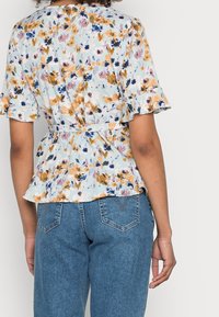 Donna che indossa una blusa floreale leggera di colore azzurro con maniche a volant e legatura in vita, abbinata a jeans in denim blu a vita alta, vista da dietro.