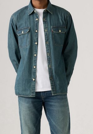 Homme portant une chemise en jean bleue boutonnée sur un t-shirt blanc avec un jean bleu, debout devant un fond clair uni.