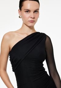 Asymmetrisk sort kjole lavet af tætsiddende stof, med en gennemskinnelig ærme og sammenkrøllede detaljer på kroppen for et struktureret udseende.