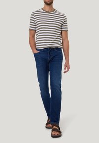 Gestreepte T-shirt met korte mouwen in olijfgroen en wit, gecombineerd met blauwe jeans en bruine sandalen. De jeans hebben een klassieke pasvorm; het T-shirt heeft een ronde hals.