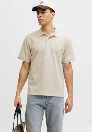 Mann iført beige poloshirt, lyseblå jeans og beige caps, holder en beige veske med svarte håndtak mot en ensfarget bakgrunn.