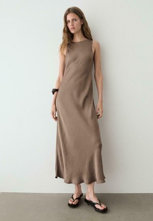 Frau trägt ein ärmelloses, knöchellanges braunes Kleid mit lockerem Schnitt, schwarzen Armreif und schwarze Sandalen mit Absatz, steht vor einer schlichten Wand.