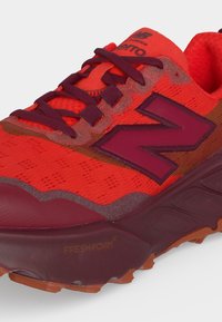 Κόκκινο και μπορντό αθλητικό παπούτσι New Balance με απορρόφηση κραδασμών Fresh Foam και υφασμάτινο επάνω μέρος με κορδόνια και μεγάλο λογότυπο "N" στο πλάι.