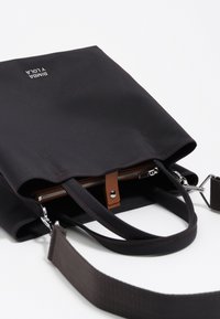 Zwarte stoffen tote bag met een ritssluiting, bruine leren accenten en zilveren hardware. Voorzien van dubbele handvatten en een afneembare schouderriem.