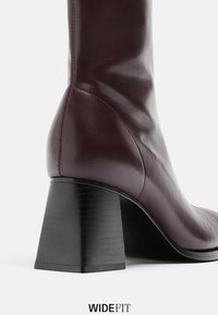 Stivaletto alla caviglia in pelle bordeaux con una forma sagomata, tacco a blocco e texture liscia. Presenta cuciture visibili e un design a vestibilità ampia.
