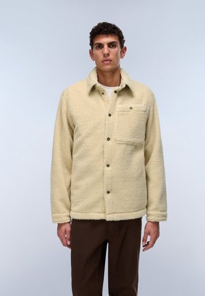 Mann trägt eine beige Fleecejacke mit Knöpfen und Brusttasche sowie dunkelbraune Hose, steht vor hellem, schlichtem Hintergrund.