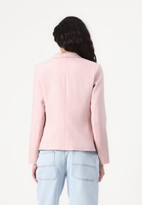 Růžový upravený blazer s klopovým límcem, dlouhými rukávy a hladkou texturou. Viditeln�ý detail švu vzadu, v kombinaci s světle modrými džíny.