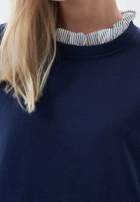 Felpa blu navy con scollatura a coste, dotata di un colletto volant a righe bianche. Texture morbida, design casual con aspetto a strati.