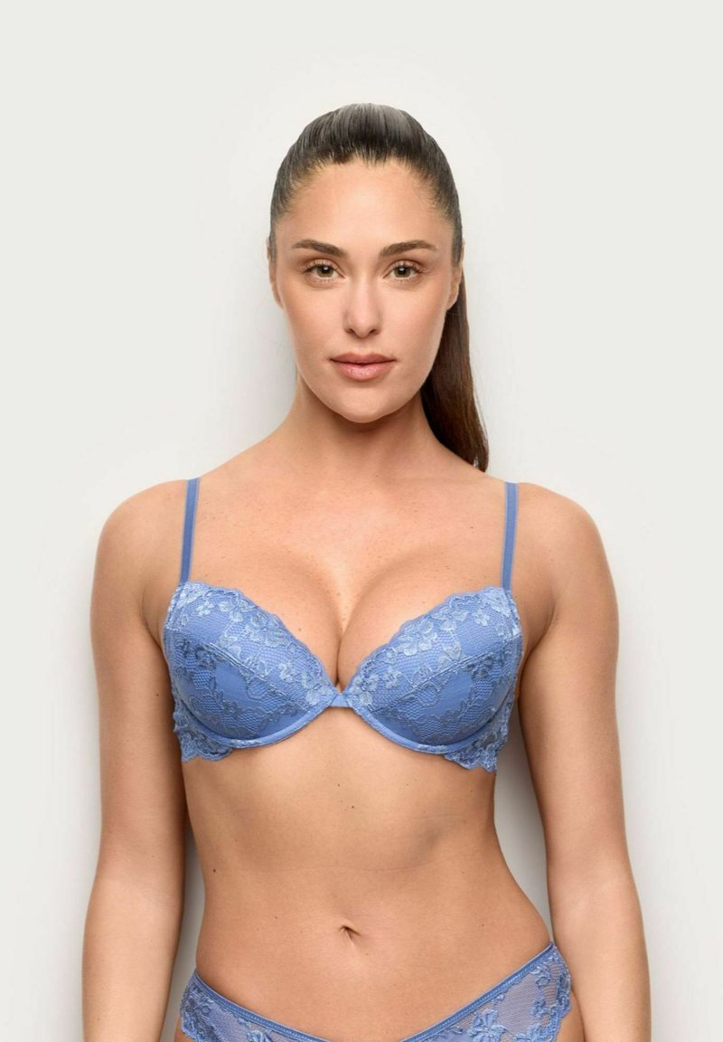 Vrouw met lang donker haar in een blauw kanten beha en bijpassende slip, staand tegen een effen lichte achtergrond, recht vooruit kijkend.