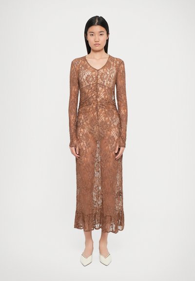 Braunes Spitzenkleid mit langen Ärmeln, einem V-Ausschnitt und einem Rüschensaum, das eine figurbetonte Silhouette und Blumenmuster aufweist. Weiße spitze Schuhe.