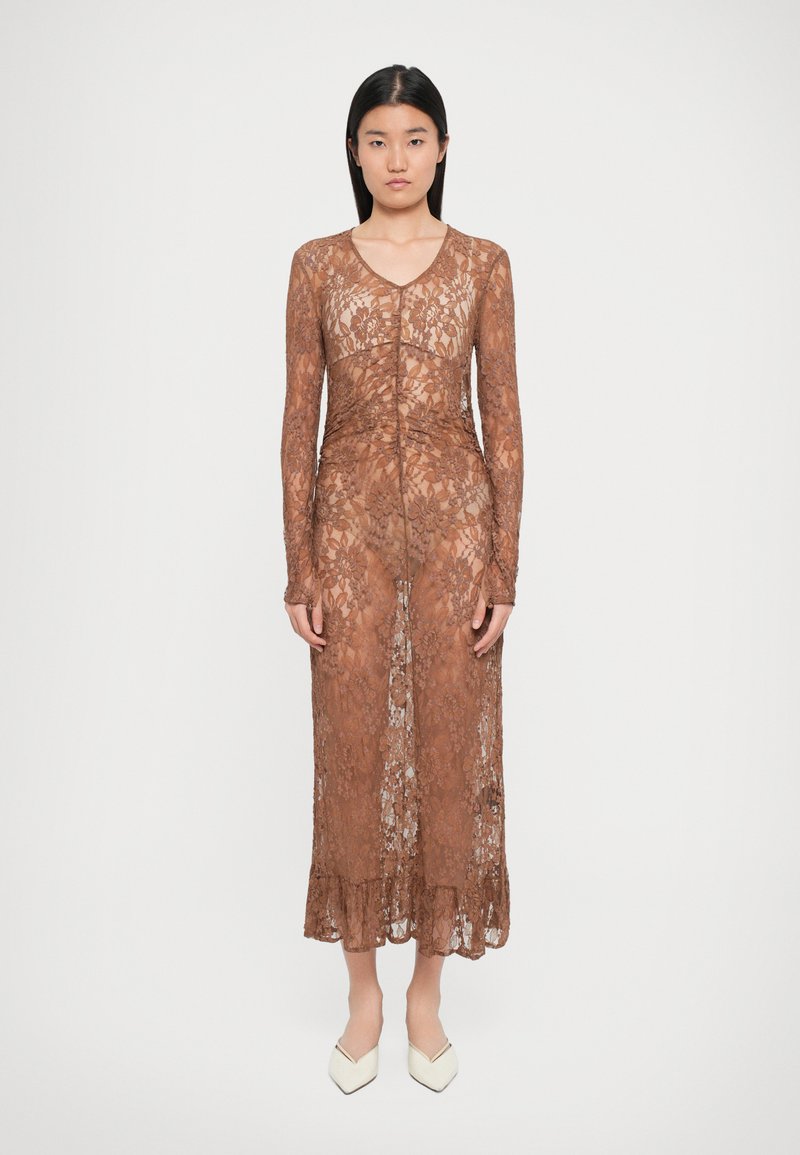 Robe en dentelle marron à manches longues, avec un décolleté en V et un ourlet à volants, présentant une silhouette ajustée et des motifs floraux. Chaussures blanches à bout pointu.