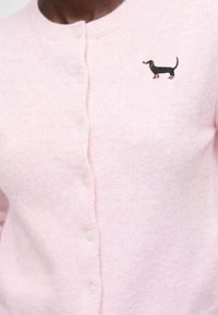 Cardigan rose clair boutonné avec un petit chien teckel noir et marron brodé sur la zone gauche de la poitrine.
