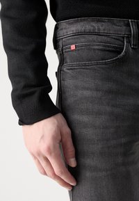 Main gauche posée à côté d'un jean en denim noir avec une étiquette rouge Hugo et un haut noir texturé à manches longues sur un fond uni.