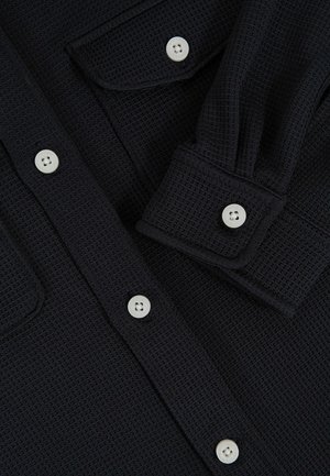 Černá texturovaná košile s dlouhým rukávem a bílými knoflíky na přední léze, s kapsou na hrudi a manžetou.