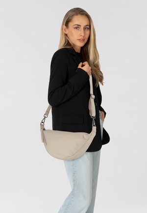 Borsa a spalla in pelle beige con forma curvata, tracolla regolabile e hardware in argento. Indossata con un blazer nero e jeans azzurri.