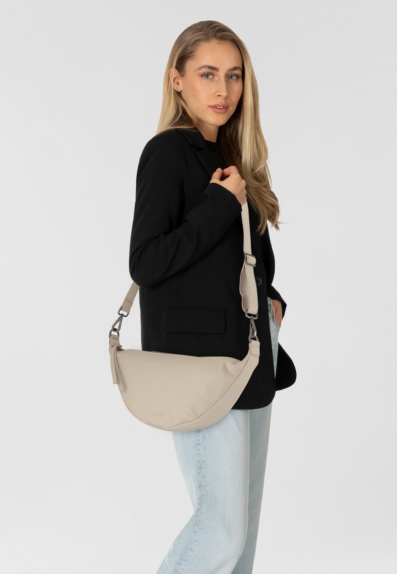 Bolso de hombro de cuero beige con forma curva, correa ajustable y herrajes plateados. Lleva puesto con un blazer negro y jeans de color azul claro.