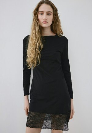 Mujer joven con cabello largo y ondulado rubio, vestida con un vestido negro de manga larga con un dobladillo de encaje negro transparente, de pie contra un fondo liso y claro.